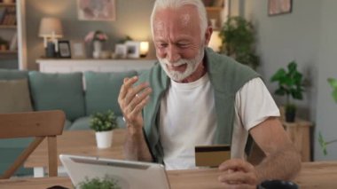 Üst düzey beyaz erkek büyükbaba evde oturuyor. İnternetten kredi kartı ve dijital tablet alışverişi yapıyor. İnternetten son sınıf öğrencisi satın almak için internet kullanıyor. 
