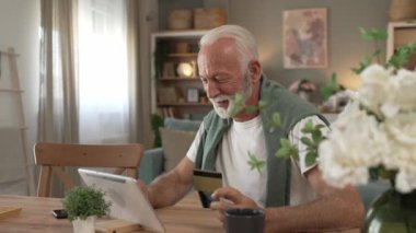 Üst düzey beyaz erkek büyükbaba evde oturuyor. İnternetten kredi kartı ve dijital tablet alışverişi yapıyor. İnternetten son sınıf öğrencisi satın almak için internet kullanıyor. 