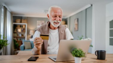 Üst düzey bir beyaz erkek büyükbaba evde oturuyor. İnternette kredi kartı ve dizüstü bilgisayar alışverişi yapıyor. İnternetten modern son sınıf öğrencilerini satın almak için internet kullanıyor.
