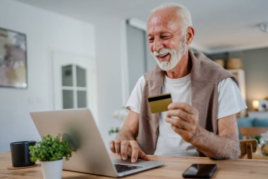 Üst düzey bir beyaz erkek büyükbaba evde oturuyor. İnternette kredi kartı ve dizüstü bilgisayar alışverişi yapıyor. İnternetten modern son sınıf öğrencilerini satın almak için internet kullanıyor.