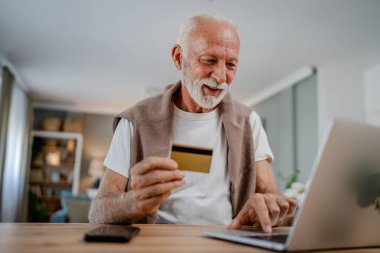 Üst düzey bir beyaz erkek büyükbaba evde oturuyor. İnternette kredi kartı ve dizüstü bilgisayar alışverişi yapıyor. İnternetten modern son sınıf öğrencilerini satın almak için internet kullanıyor.
