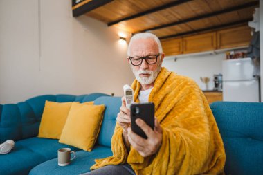 Kafkasyalı son sınıf öğrencisi kanepede oturup termometreyi tutar vücut ısısını ölçer ya da soğuk algınlığından hapşırır soğuk algınlığına yakalanır.