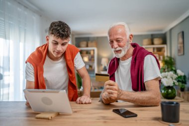 Genç bir adam ve beyaz bir erkek büyükbaba evde oturuyor. İnternet üzerinden kredi kartı ve dizüstü bilgisayar alışverişi yapıyorlar. Çevrimiçi satın almak için internet kullanıyorlar.