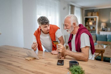 Genç bir adam ve beyaz bir erkek büyükbaba evde oturuyor. İnternet üzerinden kredi kartı ve dizüstü bilgisayar alışverişi yapıyorlar. Çevrimiçi satın almak için internet kullanıyorlar.