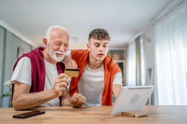 Genç bir adam ve beyaz bir erkek büyükbaba evde oturuyor. İnternet üzerinden kredi kartı ve dizüstü bilgisayar alışverişi yapıyorlar. Çevrimiçi satın almak için internet kullanıyorlar.