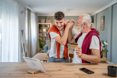 Genç bir adam ve beyaz bir erkek büyükbaba evde oturuyor. İnternet üzerinden kredi kartı ve dizüstü bilgisayar alışverişi yapıyorlar. Çevrimiçi satın almak için internet kullanıyorlar.