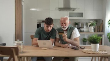 Genç bir adam ve yaşlı beyaz bir erkek büyükbaba evde otururken, çevrimiçi olarak internet üzerinden kredi kartı ve dijital tablet alışverişi yapıyorlar ve etkin modern yaşlıları satın almak için internet kullanıyorlar.