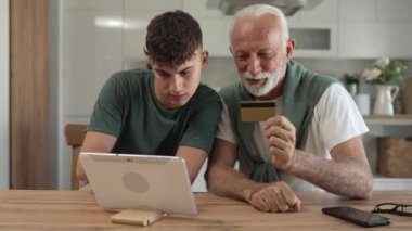 Genç bir adam ve yaşlı beyaz bir erkek büyükbaba evde otururken, çevrimiçi olarak internet üzerinden kredi kartı ve dijital tablet alışverişi yapıyorlar ve etkin modern yaşlıları satın almak için internet kullanıyorlar.