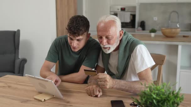 一位少年男子和一位年长的高加索男性祖父坐在家里用信用卡购物 数字平板电脑在线购物用互联网购买活跃的现代老年人 — 图库视频影像