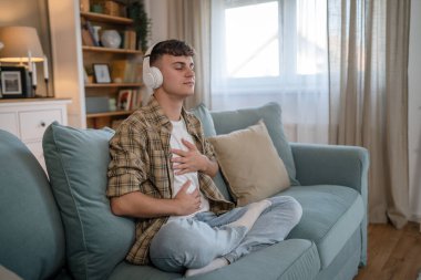 Genç bir beyaz erkek, internet rehberli meditasyon farkındalık yogası için kulaklık kullanıyor. Evde gözleri kapalı, gerçek insanlar kişisel bakım tezahürü konsepti Z jenerasyonunu taklit ediyor.