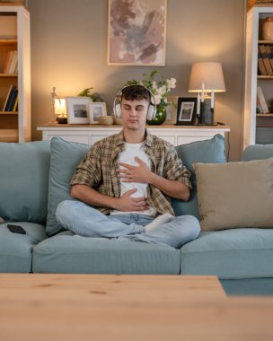 Genç bir beyaz erkek, internet rehberli meditasyon farkındalık yogası için kulaklık kullanıyor. Evde gözleri kapalı, gerçek insanlar kişisel bakım tezahürü konsepti Z jenerasyonunu taklit ediyor.