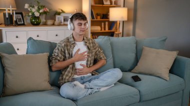 Genç bir beyaz erkek, internet rehberli meditasyon farkındalık yogası için kulaklık kullanıyor. Evde gözleri kapalı, gerçek insanlar kişisel bakım tezahürü konsepti Z jenerasyonunu taklit ediyor.