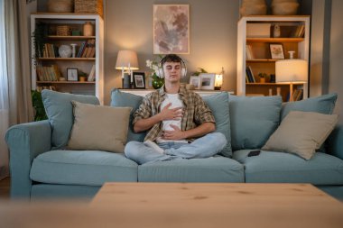 Genç bir beyaz erkek, internet rehberli meditasyon farkındalık yogası için kulaklık kullanıyor. Evde gözleri kapalı, gerçek insanlar kişisel bakım tezahürü konsepti Z jenerasyonunu taklit ediyor.