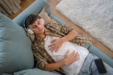 Genç bir beyaz erkek, internet rehberli meditasyon farkındalık yogası için kulaklık kullanıyor. Evde gözleri kapalı, gerçek insanlar kişisel bakım tezahürü konsepti Z jenerasyonunu taklit ediyor.