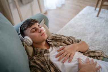 Genç bir beyaz erkek, internet rehberli meditasyon farkındalık yogası için kulaklık kullanıyor. Evde gözleri kapalı, gerçek insanlar kişisel bakım tezahürü konsepti Z jenerasyonunu taklit ediyor.