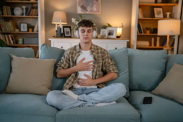 Genç bir beyaz erkek, internet rehberli meditasyon farkındalık yogası için kulaklık kullanıyor. Evde gözleri kapalı, gerçek insanlar kişisel bakım tezahürü konsepti Z jenerasyonunu taklit ediyor.