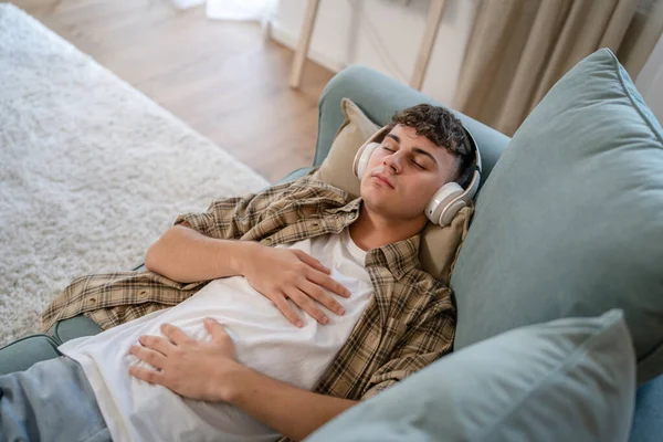 Genç bir beyaz erkek, internet rehberli meditasyon farkındalık yogası için kulaklık kullanıyor. Evde gözleri kapalı, gerçek insanlar kişisel bakım tezahürü konsepti Z jenerasyonunu taklit ediyor.