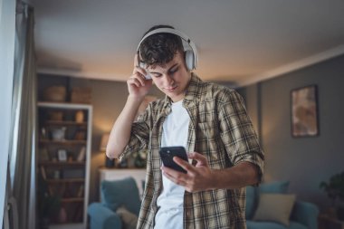 Evde dikilen bir gencin portresi müzik dinlemek ya da evde video yayını izlemek için kulaklık kullanıyor.