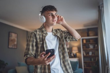 Evde dikilen bir gencin portresi müzik dinlemek ya da evde video yayını izlemek için kulaklık kullanıyor.