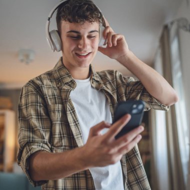 Evde dikilen bir gencin portresi müzik dinlemek ya da evde video yayını izlemek için kulaklık kullanıyor.