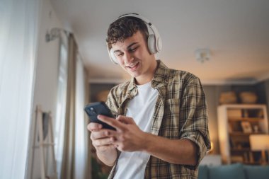 Evde dikilen bir gencin portresi müzik dinlemek ya da evde video yayını izlemek için kulaklık kullanıyor.