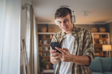 Evde dikilen bir gencin portresi müzik dinlemek ya da evde video yayını izlemek için kulaklık kullanıyor.