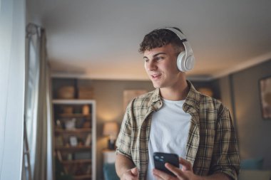 Evde dikilen bir gencin portresi müzik dinlemek ya da evde video yayını izlemek için kulaklık kullanıyor.
