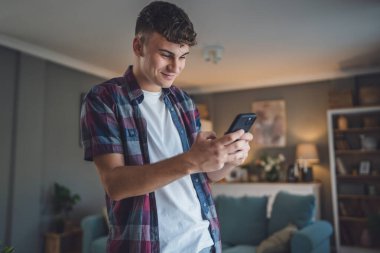 Beyaz tenli bir erkek çocuk internet üzerinden sosyal ağları taramak için akıllı cep telefonu kullanıyor ya da evde mutlu bir gülümseme mesajı atıyor.