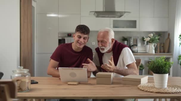 シニア男祖父年金年金の灰色の髪と孫の十代の少年とのひげは 家庭のオンラインビデオ通話の遅い動きでデジタルタブレットの前に開いた読書カード幸せな笑顔のプレゼントを受け取ります — ストック動画