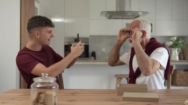 シニア男祖父年金年金の灰色の髪と彼の孫のティーンエイジャーの男の子は ホームオンラインビデオ通話の遅い動きで眼鏡で開いた幸せな笑顔でボックスにプレゼントを受け取ります — ストック動画