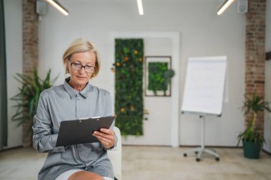 Ofiste kendine güvenen bir fotokopi odasında gözlüklü, olgun, beyaz bir kadının portresi.
