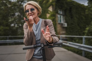 Yetişkin, beyaz, sarışın bir kadın dışarıda elektrikli tekmeyle dikiliyor scooter 'ı itiyor, cep telefonuyla telefon görüşmesi yapıyor şehirde tek başına kendine güveni tam, gerçek insanlar mekanı kopyalıyor.