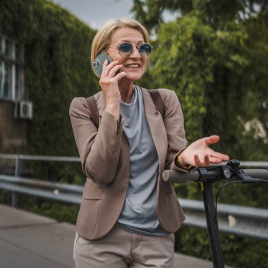 Yetişkin, beyaz, sarışın bir kadın dışarıda elektrikli tekmeyle dikiliyor scooter 'ı itiyor, cep telefonuyla telefon görüşmesi yapıyor şehirde tek başına kendine güveni tam, gerçek insanlar mekanı kopyalıyor.