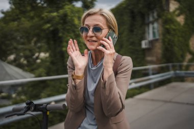Yetişkin, beyaz, sarışın bir kadın dışarıda elektrikli tekmeyle dikiliyor scooter 'ı itiyor, cep telefonuyla telefon görüşmesi yapıyor şehirde tek başına kendine güveni tam, gerçek insanlar mekanı kopyalıyor.