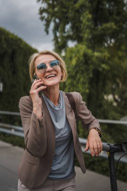 Yetişkin, beyaz, sarışın bir kadın dışarıda elektrikli tekmeyle dikiliyor scooter 'ı itiyor, cep telefonuyla telefon görüşmesi yapıyor şehirde tek başına kendine güveni tam, gerçek insanlar mekanı kopyalıyor.