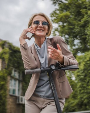 Yetişkin, beyaz, sarışın bir kadın dışarıda elektrikli tekmeyle dikiliyor scooter 'ı itiyor, cep telefonuyla telefon görüşmesi yapıyor şehirde tek başına kendine güveni tam, gerçek insanlar mekanı kopyalıyor.