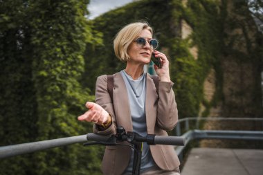 Yetişkin, beyaz, sarışın bir kadın dışarıda elektrikli tekmeyle dikiliyor scooter 'ı itiyor, cep telefonuyla telefon görüşmesi yapıyor şehirde tek başına kendine güveni tam, gerçek insanlar mekanı kopyalıyor.