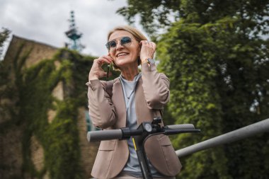 Yetişkin, beyaz, sarışın bir kadın dışarıda elektrikli tekmeyle dikiliyor scooter 'ı itiyor, cep telefonuyla telefon görüşmesi yapıyor şehirde tek başına kendine güveni tam, gerçek insanlar mekanı kopyalıyor.
