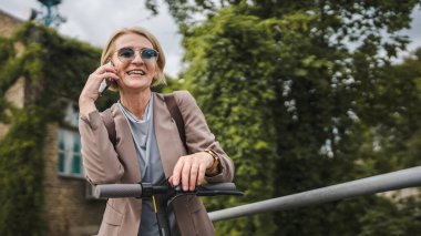 Yetişkin, beyaz, sarışın bir kadın dışarıda elektrikli tekmeyle dikiliyor scooter 'ı itiyor, cep telefonuyla telefon görüşmesi yapıyor şehirde tek başına kendine güveni tam, gerçek insanlar mekanı kopyalıyor.