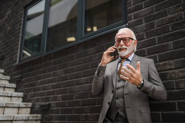 Takım elbiseli kıdemli bir işadamının portresi modern inşaat şirketinin önünde duruyor cep telefonu akıllı telefonu kullanıyor gerçek insan konuşmaları yapıyor.