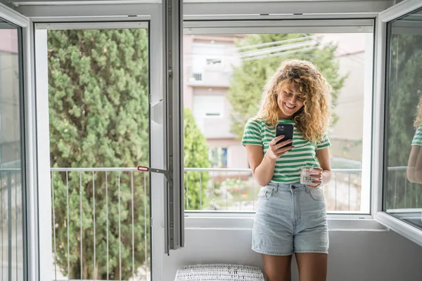 ベッドルームの自宅の窓に1人の女性の大人の慎重な女性は 携帯電話のホールドガラスを使用して 毎朝の日常的な幸せな笑顔のコピースペースをテキストメッセージを送信します — ストック写真