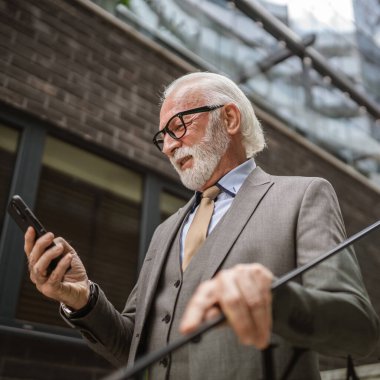 Üst düzey bir işadamı modern binanın dışında duruyor. Telefon etmek ya da mesaj atmak için cep telefonu akıllı telefonu kullanıyor. Gerçek insanlara mesaj atıyor.