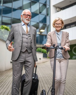 Modern bir otelin önünde bavul ve scooter ile yaşlı bir çift ve bir kadın. İş seyahati veya tatil konsepti iş adamı ve iş kadını arkadaşları veya karı koca.