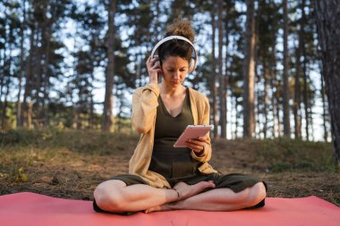 Genç yetişkin beyaz bir kadın yoga minderinde tek başına parkta ya da ormanda dijital tablet tutuyor ve kulaklıkları ayarlıyor. Online meditasyon için hazırlanıyor.