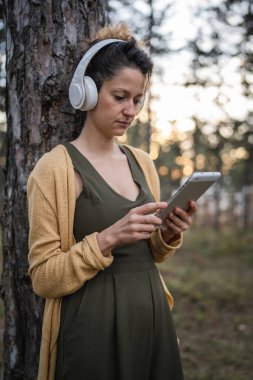 Genç bir kadın, beyaz kadın, parkta ya da ormanda kulaklıklarla, güdümlü meditasyona hazırlanıyor. Kişisel bakım, zihinsel denge kavramını uyguluyor.