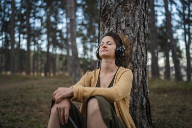 Genç yetişkin bir kadın, parkta ya da ormanda tek başına oturmuş kulaklıklarıyla meditasyona hazırlanıyor. Kişisel bakım, zihinsel denge kavramını uyguluyor.