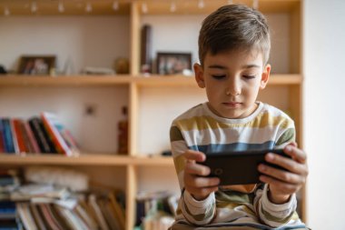 one boy caucasian child preschooler hold smartphone mobile phone at home play video games childhood and growing up technology addiction concept use smartphone app for online browsing or watch video
