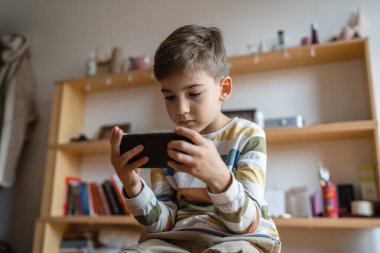 one boy caucasian child preschooler hold smartphone mobile phone at home play video games childhood and growing up technology addiction concept use smartphone app for online browsing or watch video