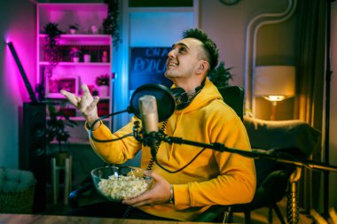 Bir beyaz erkek blogcu ya da vlogger yayın stüdyosunda video yayını yaparken el kol hareketi yapıyor mikrofon ve kulaklık kullanıyor.