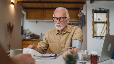 Sakallı yaşlı beyaz bir erkek evde tek başına otururken kan basıncı ölçme cihazı kullanıyor. Gerçek insanlar uzay sağlığı konseptini kopyalıyor.